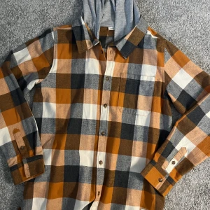 Rutig skjorta med huva från Name It - Rutig skjorta från Name It i orange, svart, grått och vitt. Skjortan har en grå huva i sweatshirtmaterial, bröstficka och knappar framtill. Tillverkad i mjuk bomull, perfekt för en chill och avslappnad stil.