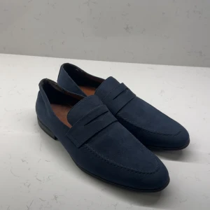 Blå mockaloafers från Iguassu Trade - Snygga loafers i mörkblå mocka från Iguassu Trade. Skorna har klassisk design med rund tå och platt sula, samt en stilren detalj över vristen. Insidan är fodrad i brunt skinn för extra komfort. Perfekta för en clean och dressad look.