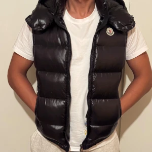 Svart dunväst från Moncler med huva - Säljer en svart, glansig dunväst från Moncler med avtagbar huva och två dragkedjefickor. Västen har quiltad design och klassisk Moncler-logga på bröstet. Perfekt för lager-på-lager och riktigt snygg till streetwear-stilen.
