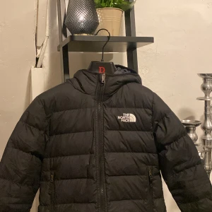 Svart dunjacka från The North Face - Säljer en svart dunjacka från The North Face, modell för barn i storlek M. Jackan har huva, dragkedja framtill och två sidofickor. Fylld med 600-fill dun för extra värme och har en klassisk, stilren look med logga på bröstet. jackan är vändbar. Passar 10-12 åringar. 