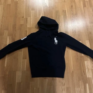 Mörkblå hoodie från Polo Ralph Lauren - Mörkblå hoodie från Polo Ralph Lauren med stor vit broderad logga på bröstet och en vit siffra 3 på ena ärmen. Riktigt stilren och passar till det mesta. Använd 3-4 gånger och köpt helt ny från ralph laurens hemsida för något år sedan. 