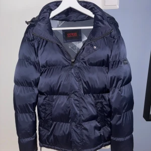 Mörkblå puffer/vinterjacka från Guess - Säljer en mörkblå puffer/vinterjacka från Guess med huva och dragkedja framtill. Jackan har en  logodetaljer på ärmen och praktiska fickor med knappar.