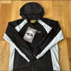 Svart och vit corteiz vindjacka  - Snygg svart och vit vindjacka från Utenos med huva och dragkedja framtill. Jackan har sportig design med kontrastpaneler och elastiska muddar vid ärmslut. Perfekt för blåsiga dagar och enkel att matcha till streetwear.