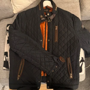 Svart quiltad jacka från Saki - Quiltad svart jacka från Saki med orange foder, flera dragkedjor och snygga bruna detaljer vid krage och fickor. Jackan har ståkrage med spänne, långärmad design och coola metalldetaljer. Perfekt för dig som gillar stilren och modern look.