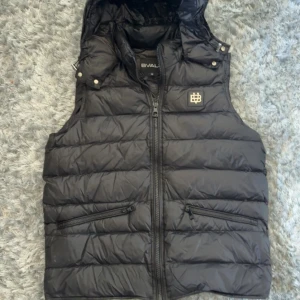 Svart dunväst från BVALDI - Snygg svart dunväst från BVALDI med dragkedja framtill och två fickor med dragkedja. Västen har en hög krage och avtagbar huva med tryckknappar. Märkets logga i metall på bröstet. Perfekt för lager-på-lager under kyliga dagar.