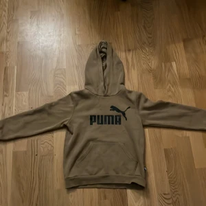 Beige hoodie från Puma med logga - Säljer en beige hoodie från Puma med svart logga framtill. Tröjan har huva, magficka och är fodrad med mjukt material på insidan. Perfekt för dig som gillar en enkel och stilren look med streetkänsla.
