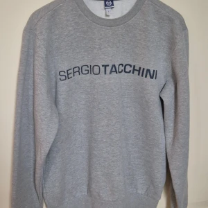 Grå sweatshirt från Sergio Tacchini L - Snygg grå sweatshirt från Sergio Tacchini med mörkblå logga tryckt över bröstet och på ärmen. Klassisk rund halsringning och ribbade muddar. Perfekt för en chill och sportig stil. Tillverkad i mjuk bomullsblandning för extra komfort.