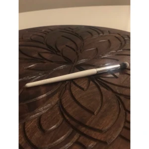 CAIA vit ögonskuggsborste E21 - Säljer en vit ögonskuggsborste från CAIA, modell E21. Borsten har ett smalt, rundat borsthuvud med mjuka syntetiska strån i svart och vitt, perfekt för att blenda och applicera ögonskugga med precision. Handtaget är stilrent och vitt med rosa CAIA-logga.