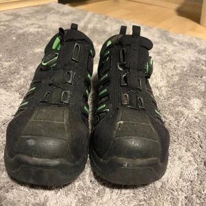 Svarta jobbskor  med gröna detaljer - Svarta jobbskor  med gröna inslag och robust design. Skorna har snörning med BOA-system, platt sula och förstärkt tå. Insidan är fodrad med ljusgrönt tyg och yttermaterialet är syntet med meshpaneler för ventilation. 