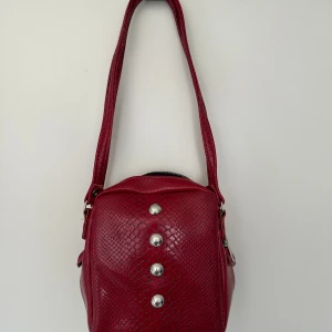 Röd axelväska med nitar och reptilmönster - Cool röd axelväska med reptilmönstrad yta och fem silvriga nitar framtill. Väskan har bred axelrem och dragkedja upptill. Insidan är fodrad med svart tyg och har en innerficka. Perfekt för dig som gillar edgy detaljer och unik design.