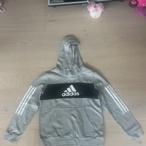 Grå Adidas hoodie med logga - Snygg original  grå oanvänd hoodie från Adidas med klassiska vita ränder på ärmarna och stor svart logga framtill. Tröjan har huva och känguruficka, perfekt för en sportig och chill stil. Mjuk insida och normal passform. Storlek 152 (11-12år)