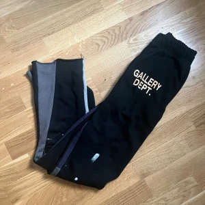 Svarta mjukisbyxor Gallery Dept. - Svarta mjukisbyxor från Gallery Dept. med tryckt logga på benet. Byxorna har elastisk midja och är tillverkade i 100% bomull. Snygga kontrastpaneler i grått längs sidorna och unika färgstänk ger en cool streetwear-vibe.
