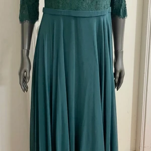 Mörkgrön långklänning med spets - Elegant mörkgrön långklänning med v-ringad överdel i spets och trekvartsärm. Kjolen är mjukt fallande och markerad i midjan med ett enkelt bälte. Perfekt för dig som vill ha en klassisk och stilren look med fina spetsdetaljer.