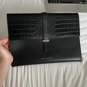 Svart datorväska från iDeal of Sweden - Stilren svart datorväska från iDeal of Sweden med croco-detaljer på locket och guldfärgad stängning. Väskan har ett elegant, minimalistiskt uttryck. Perfekt för att bära din laptop och viktiga papper.