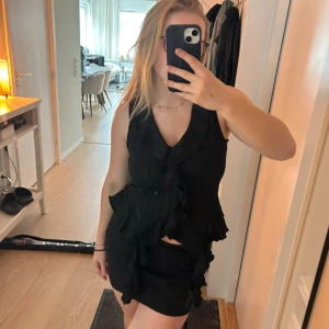 Svart topp med omlottkänsla - Supersnygg svart topp med volangdetaljer och omlottkänsla framtill. Ärmlös modell med lätt struktur i tyget och figurnära passform. Perfekt för dig som gillar trendiga och feminina plagg. Storlek M men passar en S oxå.