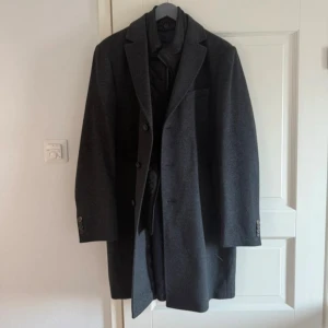 Grå rock från Sand Copenhagen - Snygg grå rock från Sand Copenhagen med klassisk krage och knäppning framtill. Insidan har ett mönstrat foder i mörkblått och en quiltad innerdel med dragkedja. Perfekt för dig som gillar stilrena och tidlösa plagg med extra detaljer.