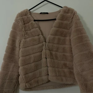 Beige pälsjacka från Gina Tricot - Säljer en fluffig och mjuk beige pälsjacka från Gina Tricot i storlek XS. Jackan har långärmade ärmar och är croppad med horisontella sömmar som ger en cool struktur. Perfekt för dig som gillar trendiga och mysiga plagg.