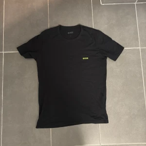 Svart t-shirt från BOSS med logga - Svart t-shirt från BOSS i klassisk modell med rund hals och korta ärmar. Diskret gul BOSS-logga på bröstet ger en clean och stilren vibe. Tillverkad i mjuk bomull för skön känsla hela dagen.