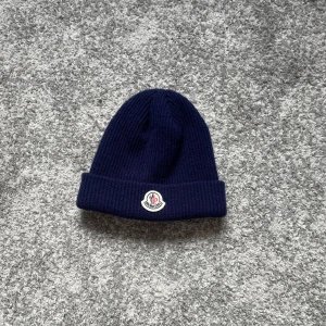 Mörkblå ribbad mössa från Moncler - Snygg mörkblå ribbstickad mössa från Moncler med uppvikt kant och klassisk logotyp framtill. Perfekt för kalla dagar och ger en clean look till din outfit. Materialet är mjukt och värmande, och färgen är riktigt djup marinblå.