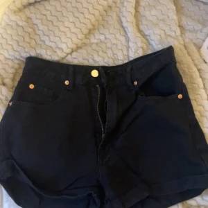 Svarta högmidjade jeansshorts - Svarta jeansshorts med hög midja och klassisk femficksdesign. De har uppvikta benslut, bälteshällor och en guldfärgad knapp framtill. Materialet är stadigt denim i bomull och de har dragkedja. Perfekta för dig som gillar en enkel och clean look.