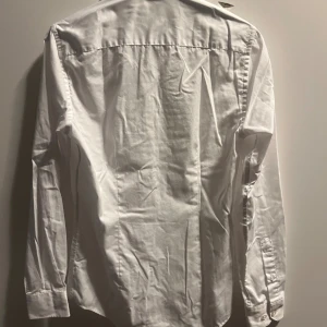 Vit klassisk skjorta i bomull - En stilren vit skjorta med lång ärm och klassisk krage. Skjortan är tillverkad i bomull och har knappar vid ärmsluten. Perfekt för en clean och enkel look. Passformen är slim fit