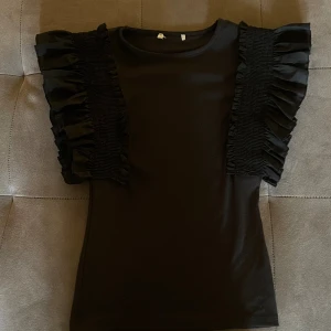 Svart topp med volangärmar - Snygg svart topp med korta ärmar och breda volangdetaljer längs axlarna. Modellen är figurnära och har rund halsringning. Perfekt för dig som vill ha en enkel men ändå trendig look. PRISET GÅR ATT DISSEKERA!!
