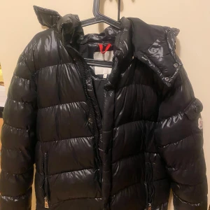Svart Moncler pufferjacka med huva - Säljer en svart pufferjacka från Moncler med glansig finish och avtagbar huva. Jackan har dragkedja framtill, två fickor med dragkedja och en ärmficka med Moncler-logga. Insidan har coolt serietryck och flera innerfickor. Perfekt för kalla dagar!