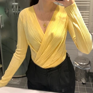 Gul bodysuit från Gina Tricot - Snygg gul bodysuit från Gina Tricot med lång ärm och plisserad struktur. Toppen har djup v-ringning och är tillverkad i ett glansigt, stretchigt material som känns mjukt mot huden. Perfekt att styla med både jeans och kjol för en fräsch look.