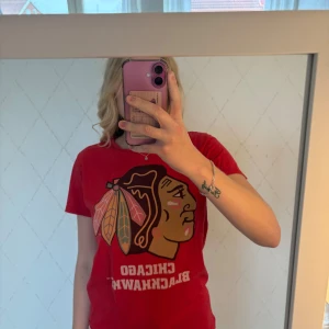 Röd Chicago Blackhawks t-shirt - Röd t-shirt med stort Chicago Blackhawks-tryck framtill. Klassisk passform med korta ärmar och rund hals. Tillverkad i mjuk bomull, perfekt för dig som gillar sportiga och snygga plagg med tydlig teamkänsla.