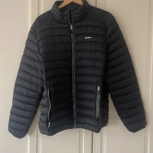 Svart pufferjacka från Tuxer - Säljer en svart pufferjacka från Tuxer med horisontella sömmar och hög krage. Jackan har dragkedja framtill och två sidofickor med dragkedja. Storlek. Är XL men sitter L