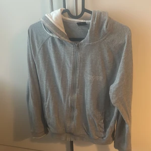 Grå hoodie från BOSS med dragkedja - Säljer en grå hoodie från BOSS med vit insida på huvan och dragkedja framtill. Tröjan har långa ärmar, fickor på sidorna och BOSS-logga på bröstet. Perfekt för chill dagar eller när du vill ha en clean look.