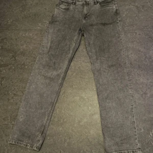 Grå jeans från Kappahl, storlek 170 - Snygga grå jeans från Kappahl i rak modell med klassisk femficksdesign. Jeansen har en tvättad look och är tillverkade i jeansmaterial med dragkedja och knapp framtill. Perfekt för dig som gillar en enkel och stilren vibe.