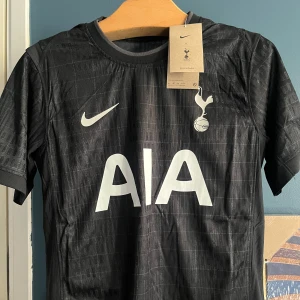 Tottenham Hotspur svart Nike matchtröja - Snygg svart Tottenham Hotspur fotbollströja från Nike med diskret rutmönster och klubbens emblem på bröstet. Tröjan har korta ärmar, rund hals och är tillverkad i ett lätt, ventilerande material. Stor AIA-logga framtill i vitt.