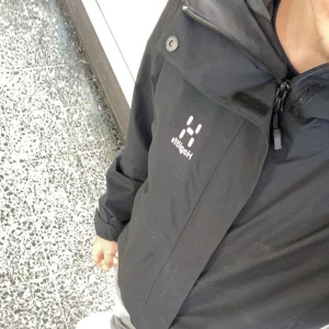 Svart vindjacka från Haglöfs - Säljer en svart vindjacka från Haglöfs med dragkedja och hög krage. Jackan har minimalistisk design med diskret logga på bröstet och praktiska detaljer som knapp vid kragen. Perfekt för dig som gillar stilren och funktionell ytterjacka.