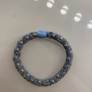 Blått armband från Kknekki - Säljer ett snyggt blått armband från Kknekki med guldiga detaljer i ett tvinnat mönster. Armbandet är gjort i ett mjukt material och har en rund form med en liten blå plastdetalj där märket står. Perfekt accessoar för att lyfta din stil.