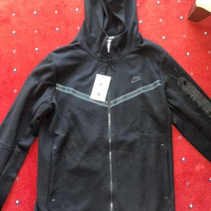 Svart Nike hoodie med dragkedja - Svart hoodie från Nike med hel dragkedja framtill, huva och diskret Nike-logga på bröstet. Snygg detalj med grått band över bröstet och ficka med dragkedja på ena ärmen. Perfekt för en sportig och avslappnad stil.