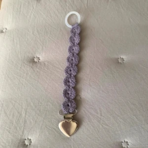 Napphållare till barn  - Bilderna visar en handgjord napphållare med lila virkad band och ett hjärtformat metallklämma ✨