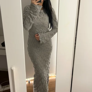 Grå långklänning - Supersnygg grå långklänning med unik strukturerad yta och långärmad design. Klänningen har en figurnära passform och coolt mönster i tyget som ger en modern vibe. Perfekt för dig som vill sticka ut med stilren look. 