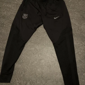 Svarta Nike FCB träningsbyxor slim fit - Svarta träningsbyxor från Nike med FC Barcelona-logga på ena benet och Nike Swoosh på andra. Slim fit-modell med snörning i midjan och Dri-Fit-material som andas. Snygga detaljer på benen och perfekt passform för fotboll eller träning.