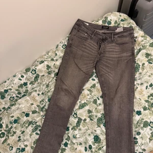Grå jeans från Jack & Jones - Snygga grå jeans från Jack & Jones med klassisk femficksdesign och svarta detaljer på bakfickan. Jeansen har en rak passform och är tillverkade i mjukt denimtyg som sitter skönt hela dagen. Perfekt för dig som gillar stilrena och enkla jeans.