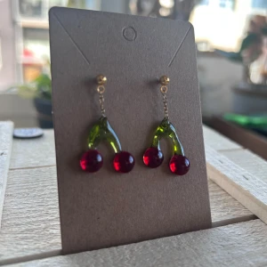 Körsbärsörhängen i glas-look - Supergulliga örhängen med körsbärsdesign! Hängande röda bär och gröna stjälkar i glasliknande material, fästa på små guldfärgade pluppar. Perfekt för dig som vill sticka ut med en lekfull och färgglad accessoar.