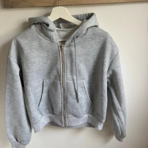 Grå hoodie med dragkedja och huva - Säljer en grå hoodie i storlek S. Tröjan har huva med snörning, hel dragkedja framtill och två stora fickor. Tillverkad i mjuk polyester, perfekt för chill dagar. Ärmarna är långärmade och muddade vid avslut.