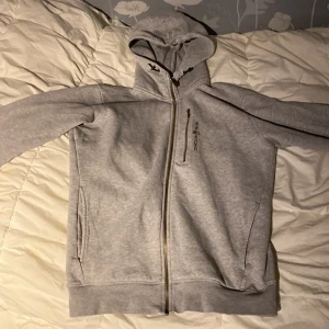 Grå hoodie från Sail racing  - Säljer en grå hoodie från Sail racing med hel dragkedja och snörning i huvan. Tröjan har en bröstficka med dragkedja och två sidofickor. Mjuk och skön bomullsmix, perfekt för chill eller träning. Långärmad och med klassisk passform. Defekt är att det är lite svart/grå färg på ryggen och lite på framsidan men ingen man riktigt lägger märke till vid första glimts 