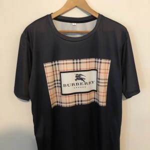 Mörkblå Burberry t-shirt med rutor - Snygg mörkblå t-shirt från Burberry med klassiskt rutigt mönster och logga på bröstet. T-shirten har rund halsringning och korta ärmar. Materialet känns mjukt och lätt, perfekt för en stilren look.