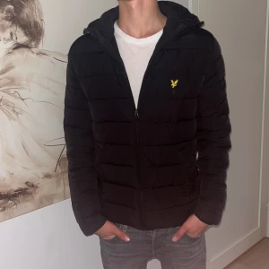 Svart pufferjacka från Lyle & Scott - Svart pufferjacka från Lyle & Scott med huva och dragkedja framtill. Jackan har det klassiska gula logobroderiet på bröstet och quiltad design. Perfekt för kyliga dagar och har en clean, stilren look. Modellen är 182. Storleken är S skulle kunna passa M. 