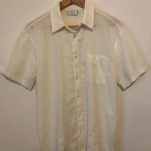 Vit linneskjorta från Grunt XL - Vit skjorta från Grunt i storlek XL, tillverkad i linne. Klassisk krage, korta ärmar och en bröstficka. Skjortan har knappar framtill och en clean, stilren look som funkar till många olika stilar.