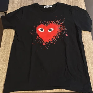 Svart t-shirt Comme des Garçons Play - Svart t-shirt från Comme des Garçons Play med ett stort rött hjärta med ögon på bröstet och coolt splattertryck runtom. Klassisk rund hals och korta ärmar. Materialet är mjuk bomull som känns skönt mot huden. Perfekt statement-plagg för dig som gillar lite mer soft style .