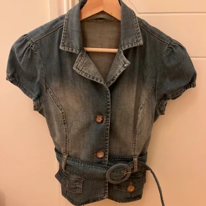 Kort jeansjacka med bälte och puffärm - Snygg kort jeansjacka från Dollhouse med puffärmar, markerad midja och brett bälte med rund spänne. Jackan har knappar framtill, fickor och klassisk krage. Perfekt för dig som vill ha en unik denimlook med coola detaljer.