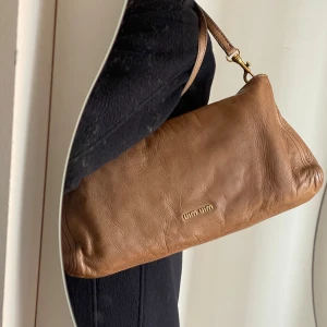 Vintage Miu Miu Axelväska  - Snygg brun axelväska från Miu Miu i mjukt italienskt skinn med justerbar rem och guldfärgade metalldetaljer. Väskan har minimalistisk design med diskret logga framtill och dragkedja upptill. Perfekt för dig som gillar stilrena accessoarer.