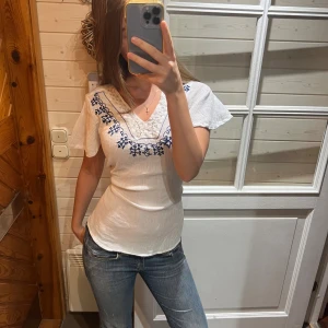 Vit blus med blå broderi från Fashion Quality - Säljer en vit blus från Fashion Quality i storlek 42/44. Blusen har korta, luftiga ärmar och v-ringad hals med blå broderade detaljer längs ringningen. Materialet är lätt och känns som bomull, perfekt för vår och sommar. Snygg och chill stil med fina broderier.